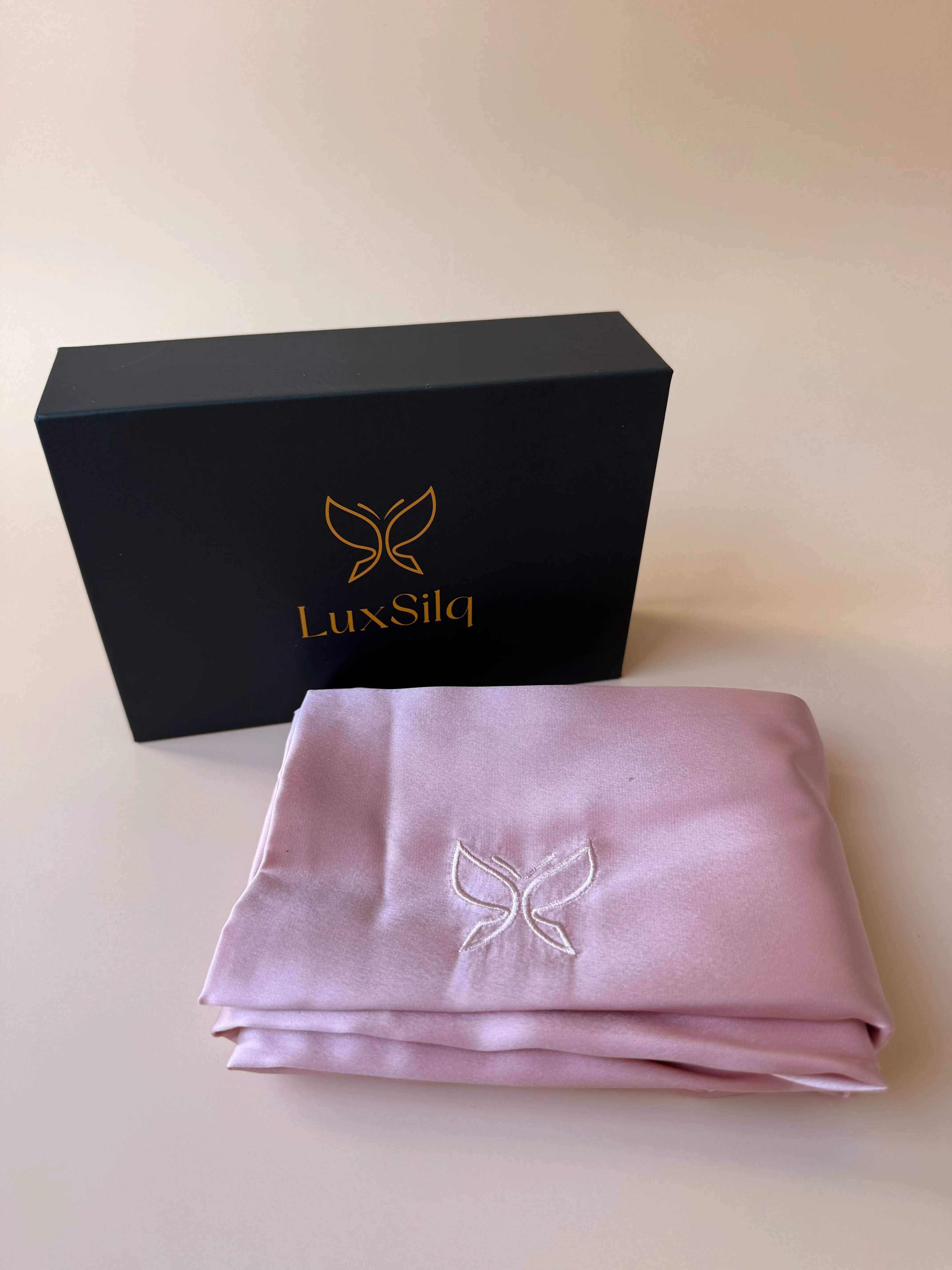 Folded pink fabric with embroidered butterfly design next to a black box with 'LuxSilq' branding on a light 
blush pinkbackground.